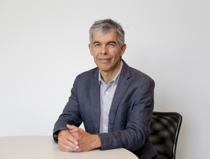 El profesor Osvaldo Larrañaga, sentado con sus manos apoyadas en una mesa. Fotografía: Banco de Imágenes UC.