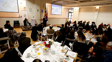 Grupo de personas sentadas en una mesa con café y galletas, escuchando al rector Ignacio Sánchez. Fotografía: César Cortés