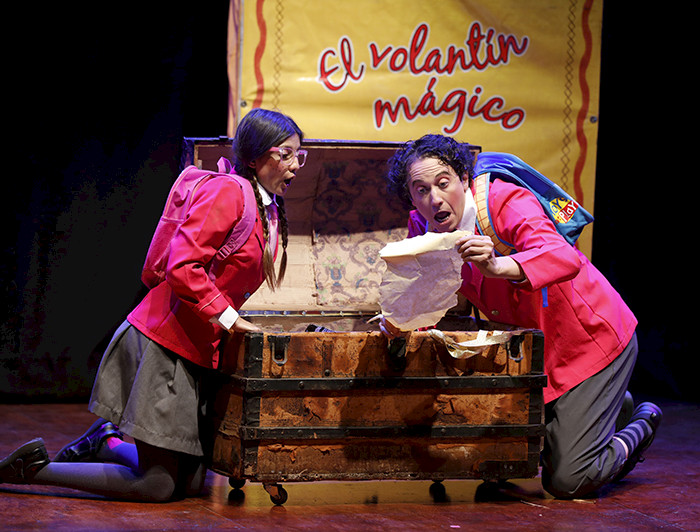 Rosario y Gaspar, dos escolares, sentados frente a un baúl de madera, descubriendo un objeto antiguo. Detrás de ellos, un libro grande dice El Volantín Mágico.