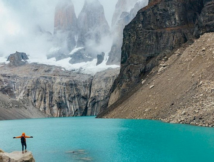 Persona con sus brazos abiertos mira el lago y al fondo las Torres del Paine.