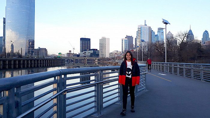 Tamara Abarca is posing for the photo on a bridge in Philadelphia. Behind her are a lot of buildings and water.
